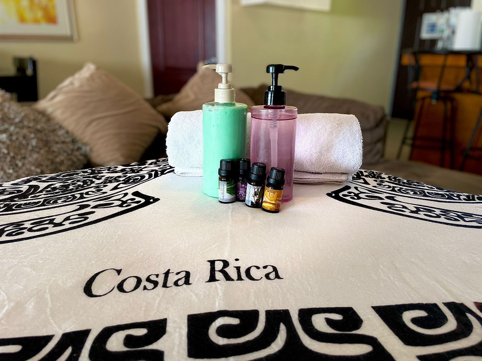 Een bed met een beddegoed met Costa Rica thema, een witte handdoek en diverse toiletartikelen, waaronder een groene fles, een roze fles en meerdere kleine flesjes essentiële oliën.