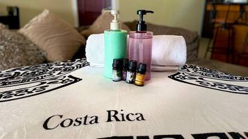 Een bed met een beddegoed met Costa Rica thema, een witte handdoek en diverse toiletartikelen, waaronder een groene fles, een roze fles en meerdere kleine flesjes essentiële oliën.