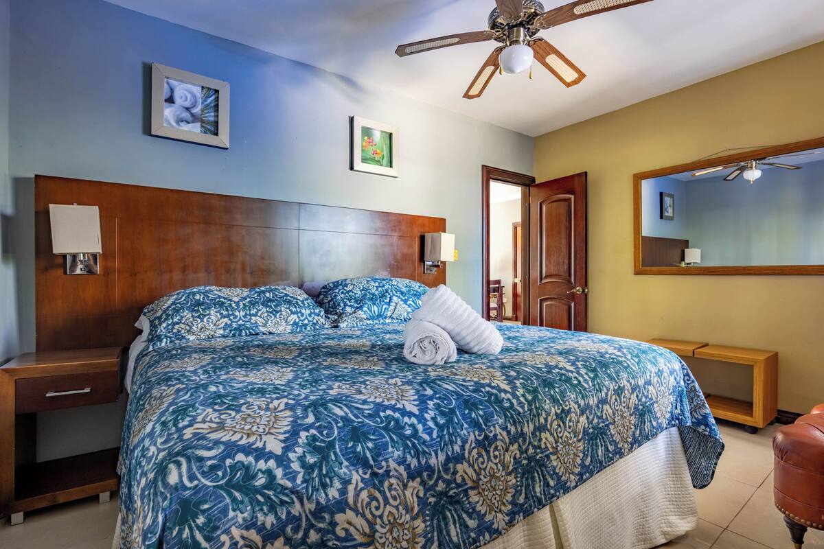 A hotel room with a large bed, wooden headboard, two bedside tables, a ceiling fan, and a mirror on the wall.