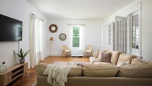 A living room with a beige sofa, two armchairs, a flat-screen TV, and a large window with curtains.