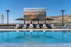 Zona de piscina con sillones de descanso y sombrillas, una pantalla grande mostrando una vista panorámica y un cielo despejado.