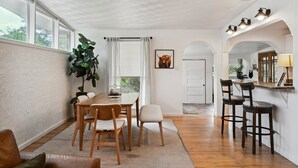 Uma área de refeições com mesa e cadeiras de madeira, uma planta grande e um espaço de cozinha com banquetas e balcão.