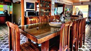 A dining area with a glass-top table surrounded by wooden chairs, a television, and a bar area with shelves of bottles. - 3 Bed House - Sleeps 20 - Pets - Pool - Garden (La Union)