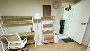 Een kleine kamer met een houten bureau, een witte stoel, een witte plank en een witte prullenbak, en een klok aan de muur.