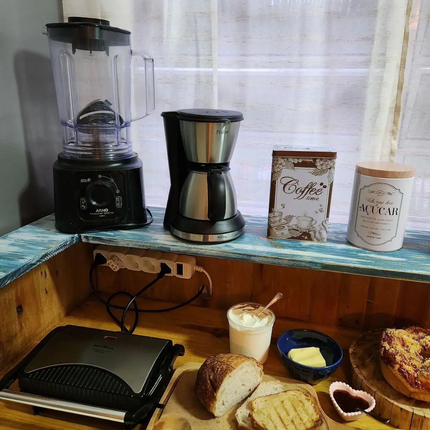 Un piano di lavoro in cucina con frullatore, macchina del caffè, contenitore per caffè, zuccheriera, tostapane, burro, pane e barattolo di marmellata.