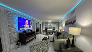 A modern living room with a flat-screen TV, a sofa, a patterned ottoman, and a dining area in the background.