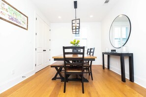 Uma sala de jantar com uma mesa de madeira, cadeiras, um vaso com flores, uma luminária pendente e um espelho na parede.