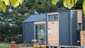 A modern, compact house with a dark exterior and wooden accents, featuring a small deck and a visible chicken in the yard. - Sugar Hut by Tiny Away (Great Horkesley)