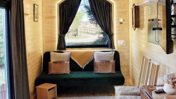 A cozy cabin room with a wooden interior, a green sofa with cushions, a small wooden table, and a window with curtains. - Sugar Hut by Tiny Away (Great Horkesley)