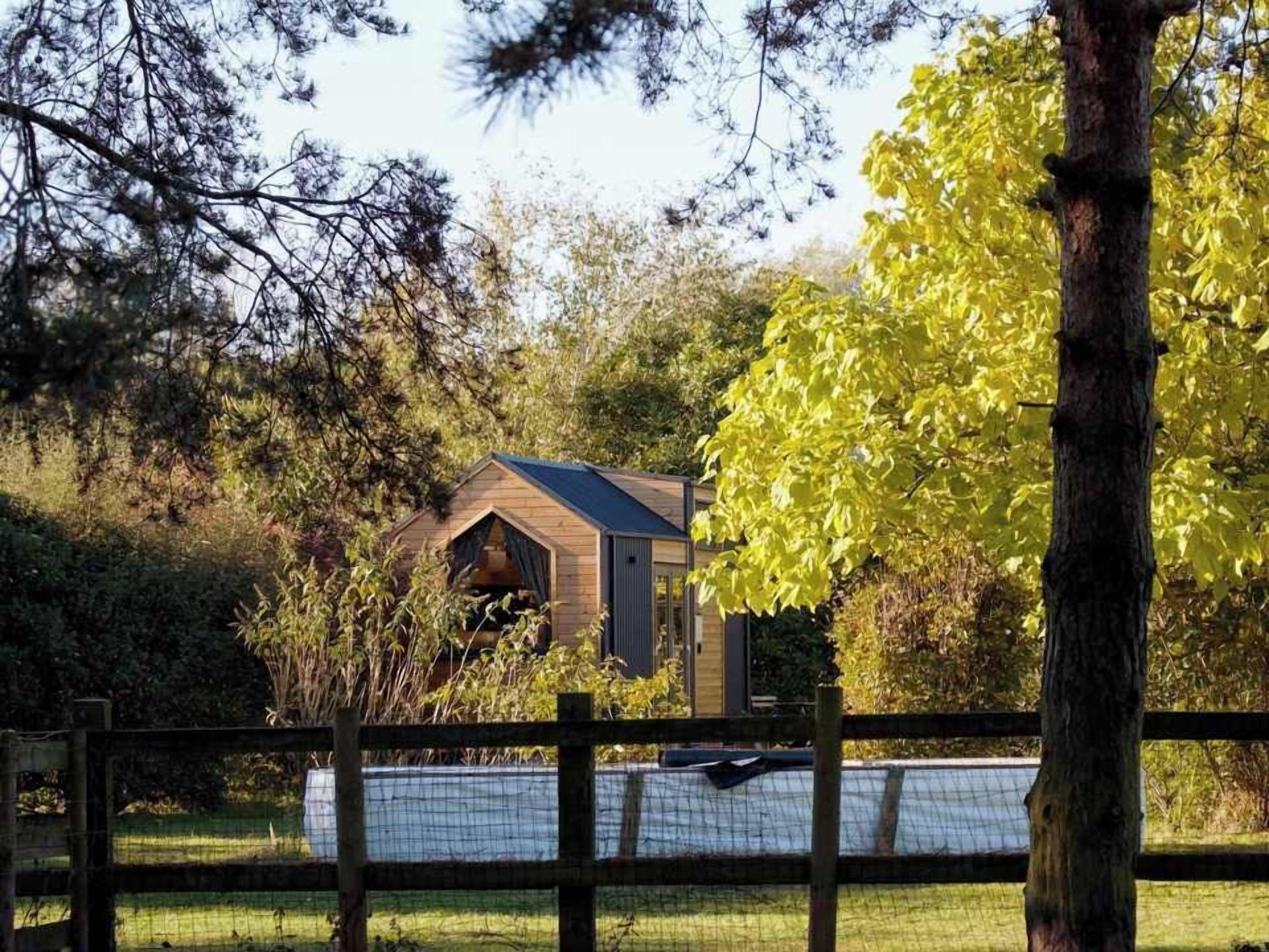 A small wooden cabin with a gabled roof, surrounded by trees and a fenced area.