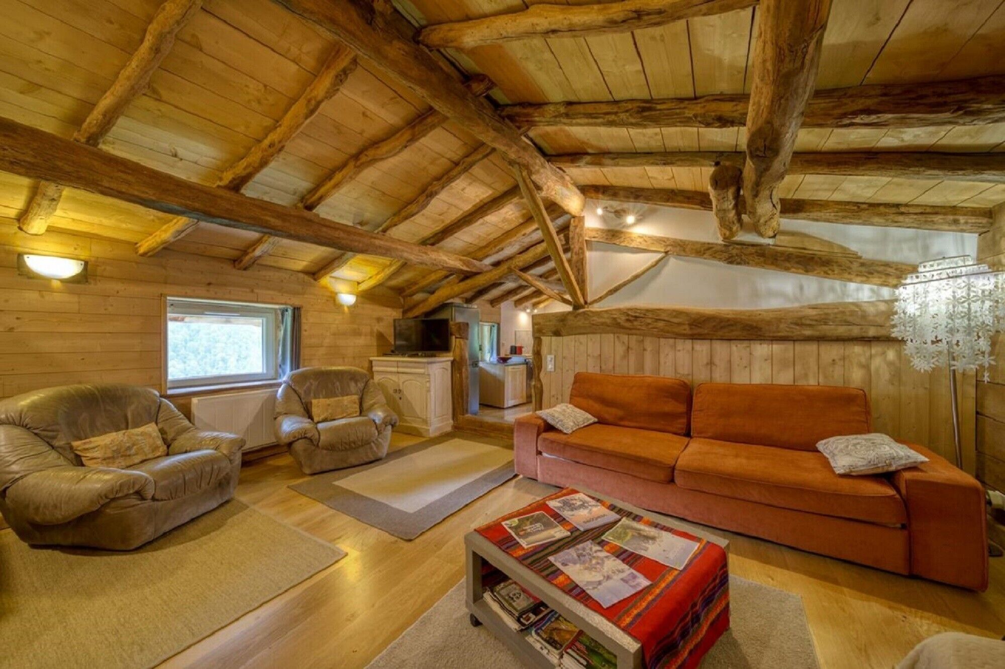 A cozy cabin interior with a wooden ceiling, leather sofas, a coffee table with magazines, and a window providing natural light.