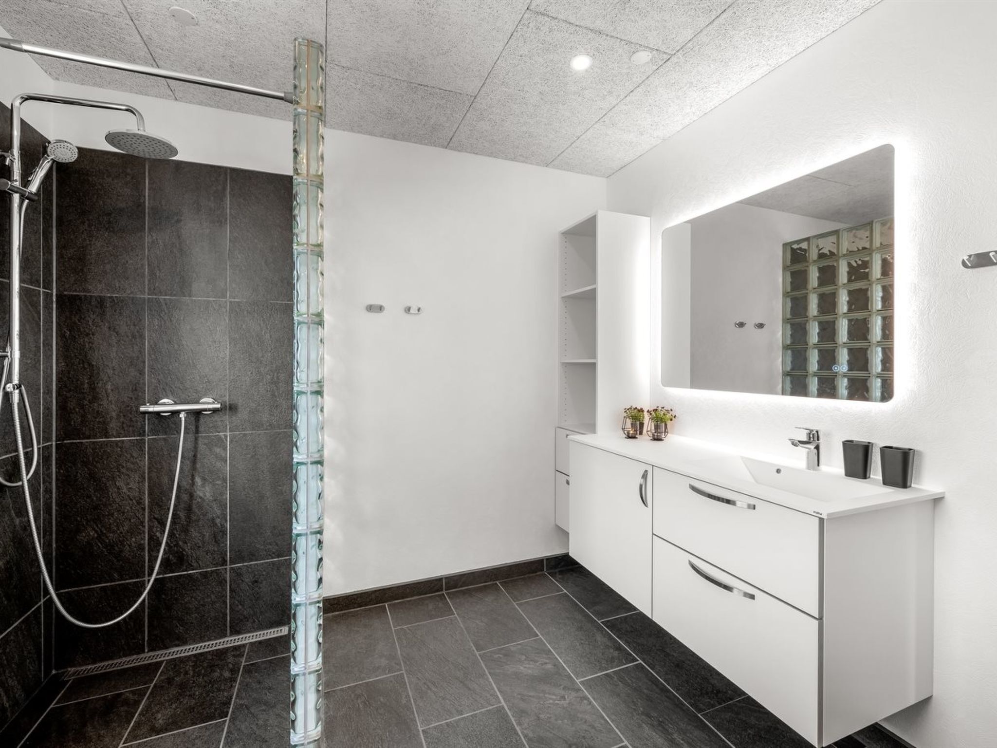 Un baño moderno con cabina de ducha de vidrio, tocador blanco con espejo y suelo de baldosas oscuras.