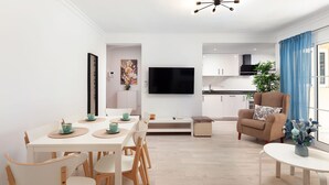 A modern living room with a dining area, a kitchen, and a TV mounted on the wall.