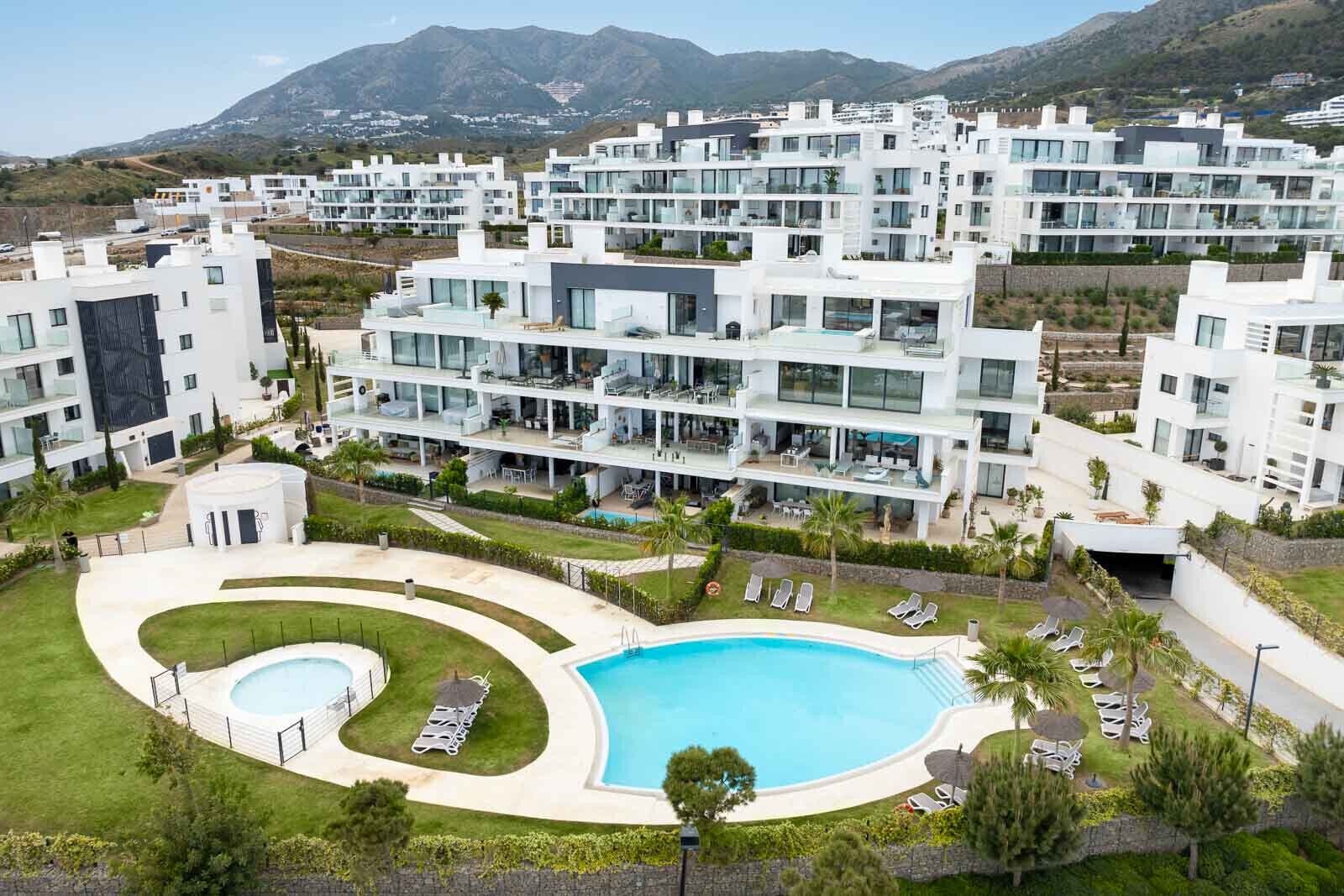 A modern residential complex with multiple white buildings, a central swimming pool, and surrounding greenery.