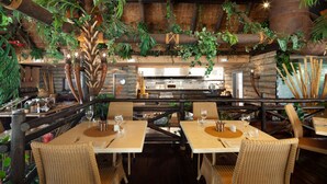 Un restaurante con mesas y sillas de madera, rodeado de plantas colgantes y una pared de ladrillo rústico.