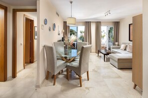 A modern living room with a glass dining table, grey upholstered chairs, a beige sofa, and a view of the outdoors.