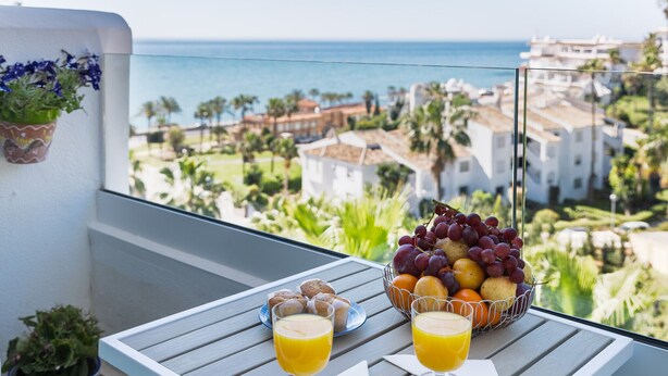 Un balcón con una mesa lista para el desayuno, un cuenco de fruta y dos vasos de zumo de naranja.