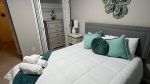 Un lit bien fait, avec une couette blanche matelassée, des oreillers turquoise et une tête de lit grise. Une table de chevet blanche, sur laquelle se trouve une lampe et un objet décoratif fixé au mur, est placée à côté du lit.