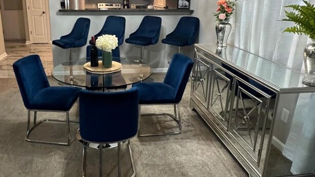 Un espace repas moderne avec une table en verre et des chaises bleues, un lustre et une cuisine en arrière-plan.