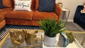 Un salon moderne avec un canapé, une table basse agrémentée d’objets décoratifs et un coussin sur lequel est inscrit : « SOYEZ LES BIENVENUS ».
