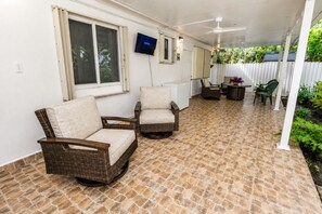 Ein überdachter Patio-Bereich mit zwei Korbstühlen, einem kleinen Tisch und einem an der Wand befestigten Fernseher.