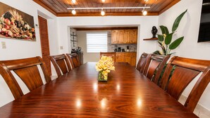 Une salle à manger avec une table et des chaises en bois, un vase de fleurs, et une cuisine en arrière-plan.