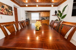 Comedor con mesa y sillas de madera, un jarrón de flores y una cocina al fondo.