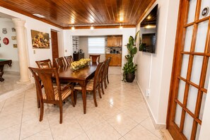 A dining area with a wooden table and chairs, a kitchen in the background, and a large potted plant. - House In The Heart of Miami (Miami)