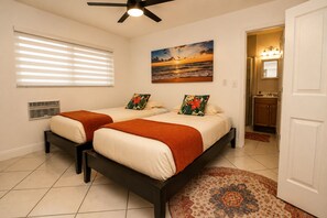 Une chambre d’hôtel avec deux lits, un ventilateur de plafond, un tableau représentant un coucher de soleil et une salle de bain avec un lavabo et un miroir.