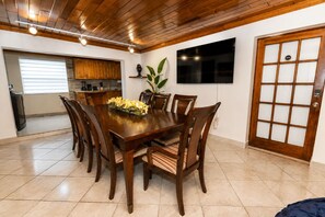 Un coin repas avec une table et des chaises en bois, un téléviseur fixé au mur et une cuisine en arrière-plan.