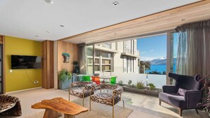 A modern living room with a wooden coffee table, two patterned chairs, a purple armchair, and a large window offering a view of a mountain and sea. - Lakeview Park Apartment (Wanaka)