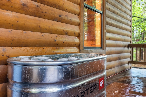 A metal barrel with condensation, placed on a wooden deck outside a log cabin with a window.