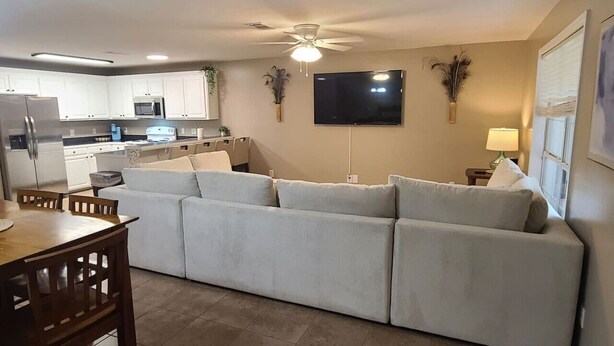 Una cocina moderna con armarios blancos, horno empotrado y refrigerador. Hay una zona de comedor con una mesa y sillas de madera. Un sofá grande se encuentra en la sala de estar, y un televisor de pantalla plana está montado en la pared.
