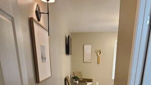 Un pasillo con paredes color beige, una puerta blanca y una lámpara. Se observa un cuadro en la pared y una pequeña mesa con una silla al fondo.