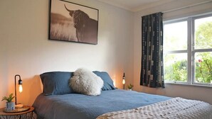 A bedroom with a large bed, a side table with a lamp, a framed picture on the wall, and a window with curtains. - Rose End Escape.
Home comfort in the central Wairarapa country side  (Dalefield)