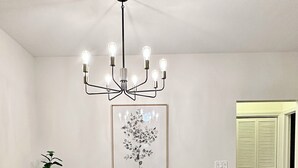 Ein Essbereich mit einem Glastisch, vier braunen Stühlen, einer schwarzen Pendelleuchte und einem gerahmten Blumenbild an der Wand.