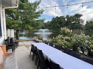 Un espace extérieur aménagé pour les repas, offrant une vue sur un lac et une fontaine, et entouré d’arbres et de plantes en pot.