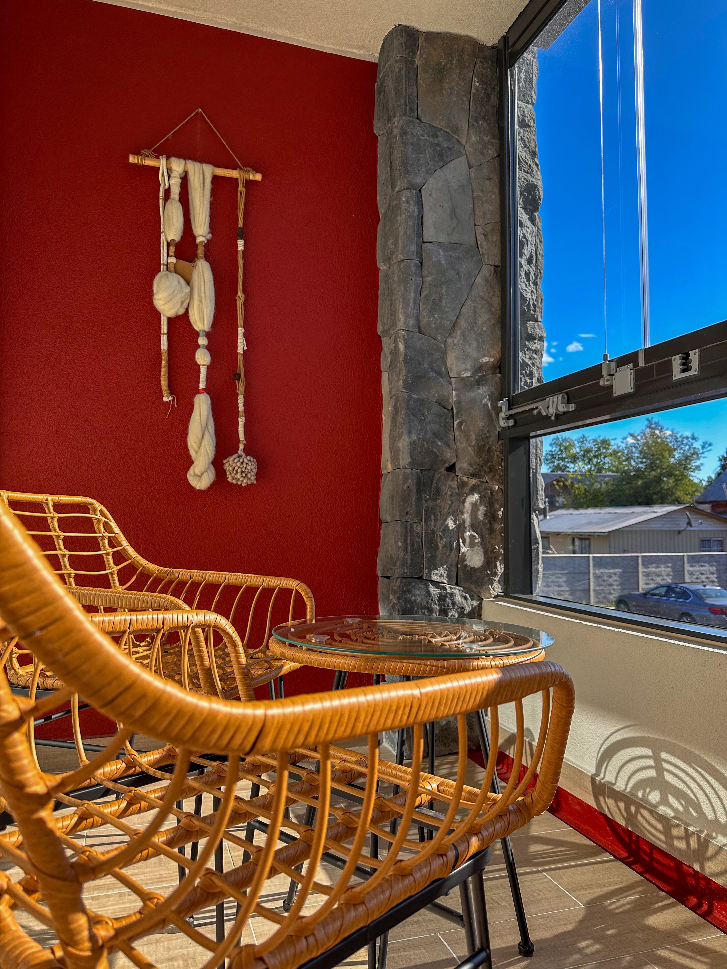 A room with wicker chairs, a glass table, a stone wall, and a view of a car through a window.