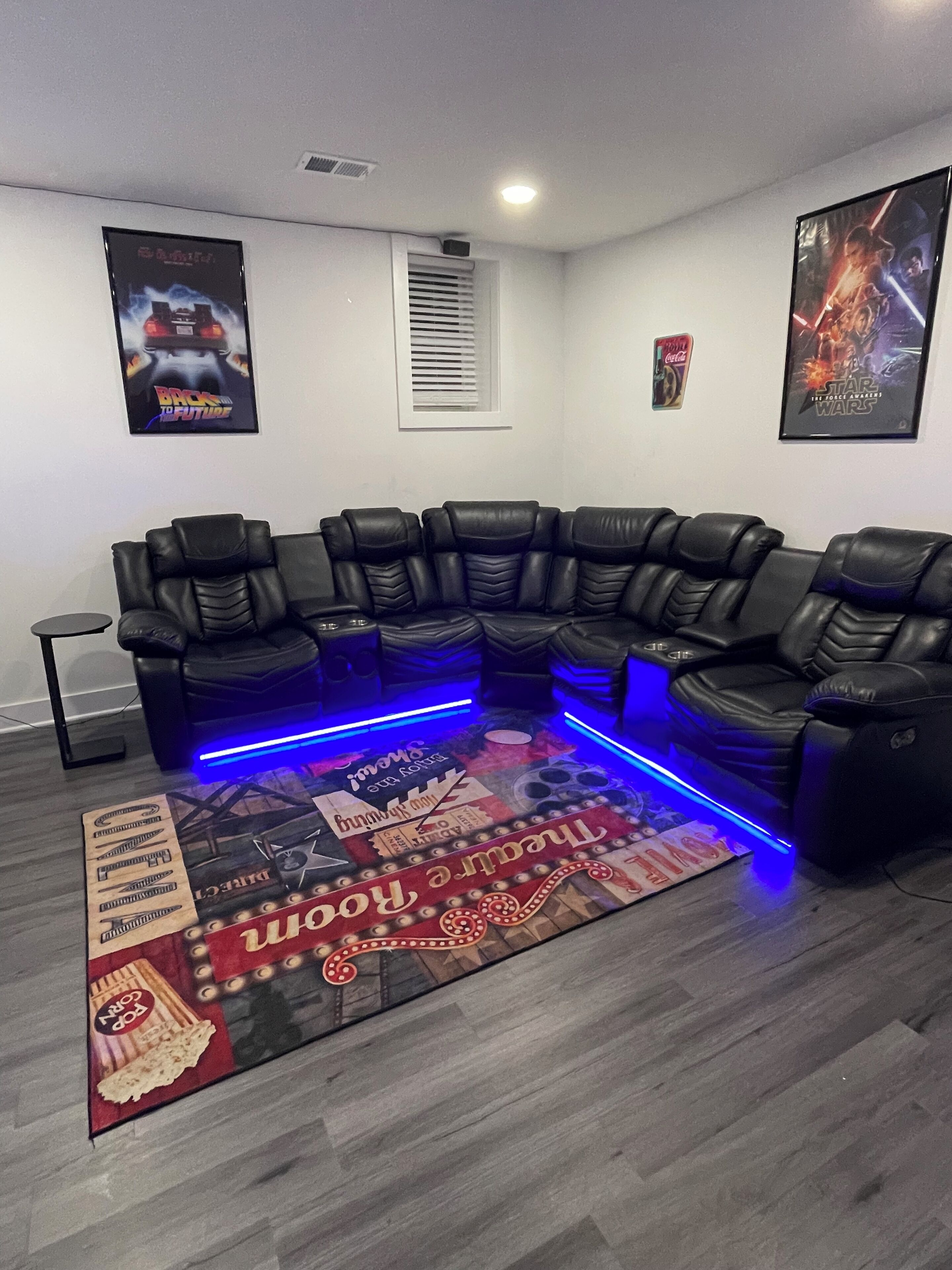 A modern living room with a black leather sectional sofa, a patterned rug, and two posters on the walls.