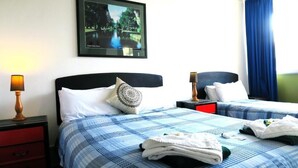 Habitación de hotel con dos camas, una mesita de noche con lámpara, un cuadro enmarcado en la pared y una ventana con cortinas azules.