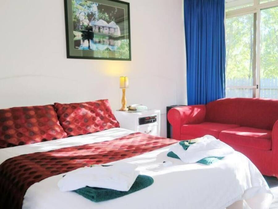 A hotel room with a bed, a red armchair, a nightstand with a lamp, and a window with a blue curtain.