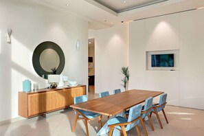A modern dining area with a wooden table and blue chairs, a wooden cabinet, a large round mirror, and a flat-screen TV mounted on the wall.