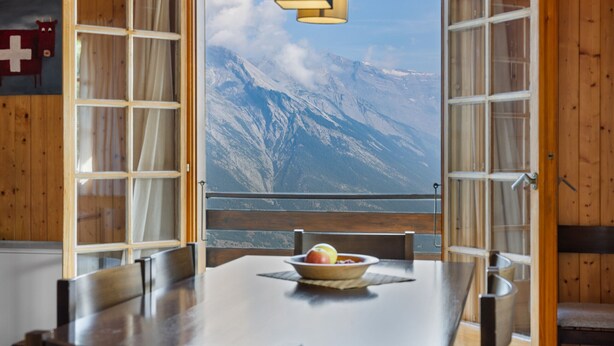 A wooden interior with a dining table and chairs, a bowl of fruit, and a view of snow-covered mountains through open windows.