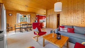 A spacious living area with a wooden ceiling, a dining table, a red chair, a grey sofa, and a kitchen in the background.