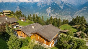 A mountain cabin with a sloped roof, surrounded by lush greenery and a view of towering mountains.