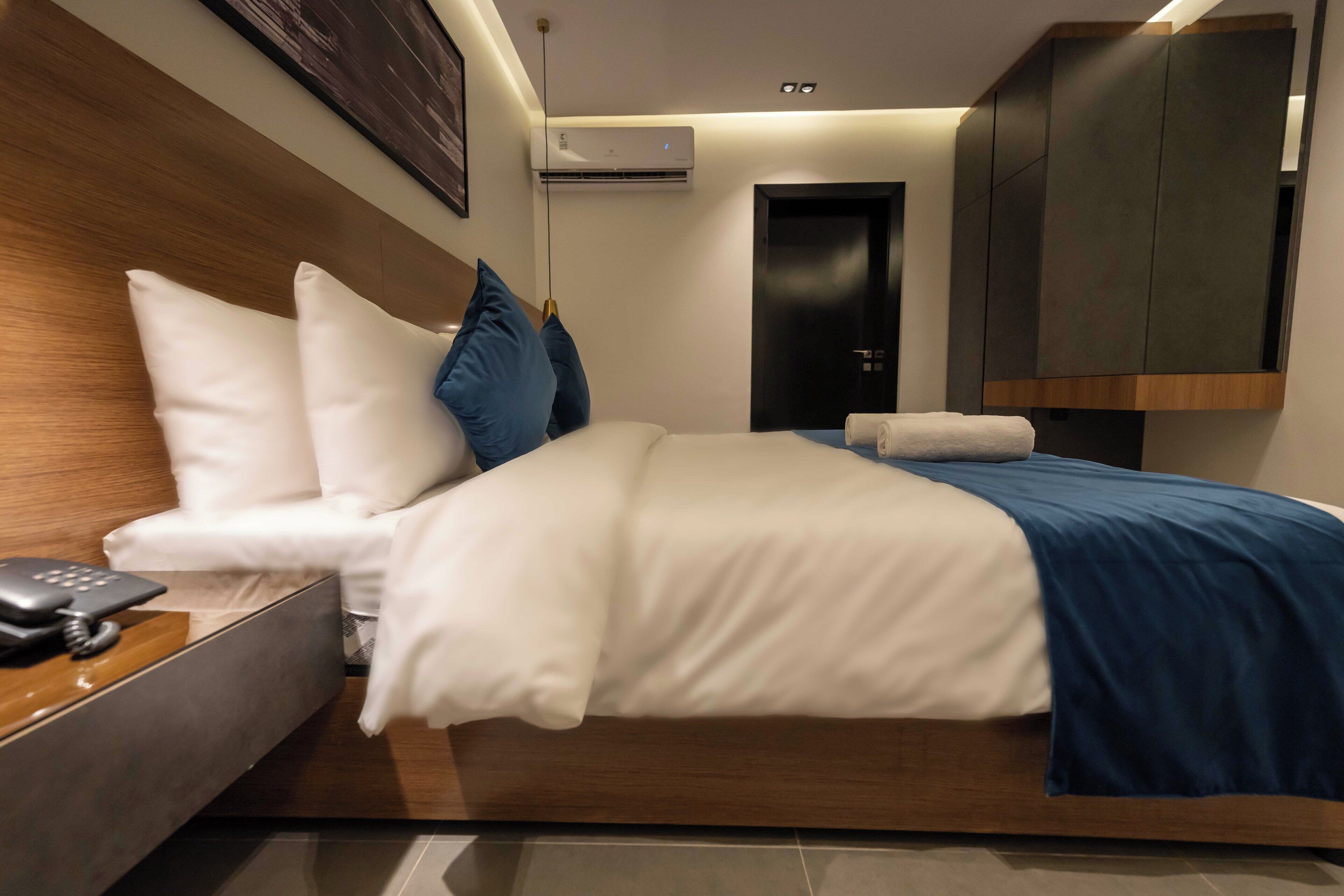 A modern hotel room with a wooden headboard, a bed with white and blue bedding, a bedside table with a phone, and a wardrobe.