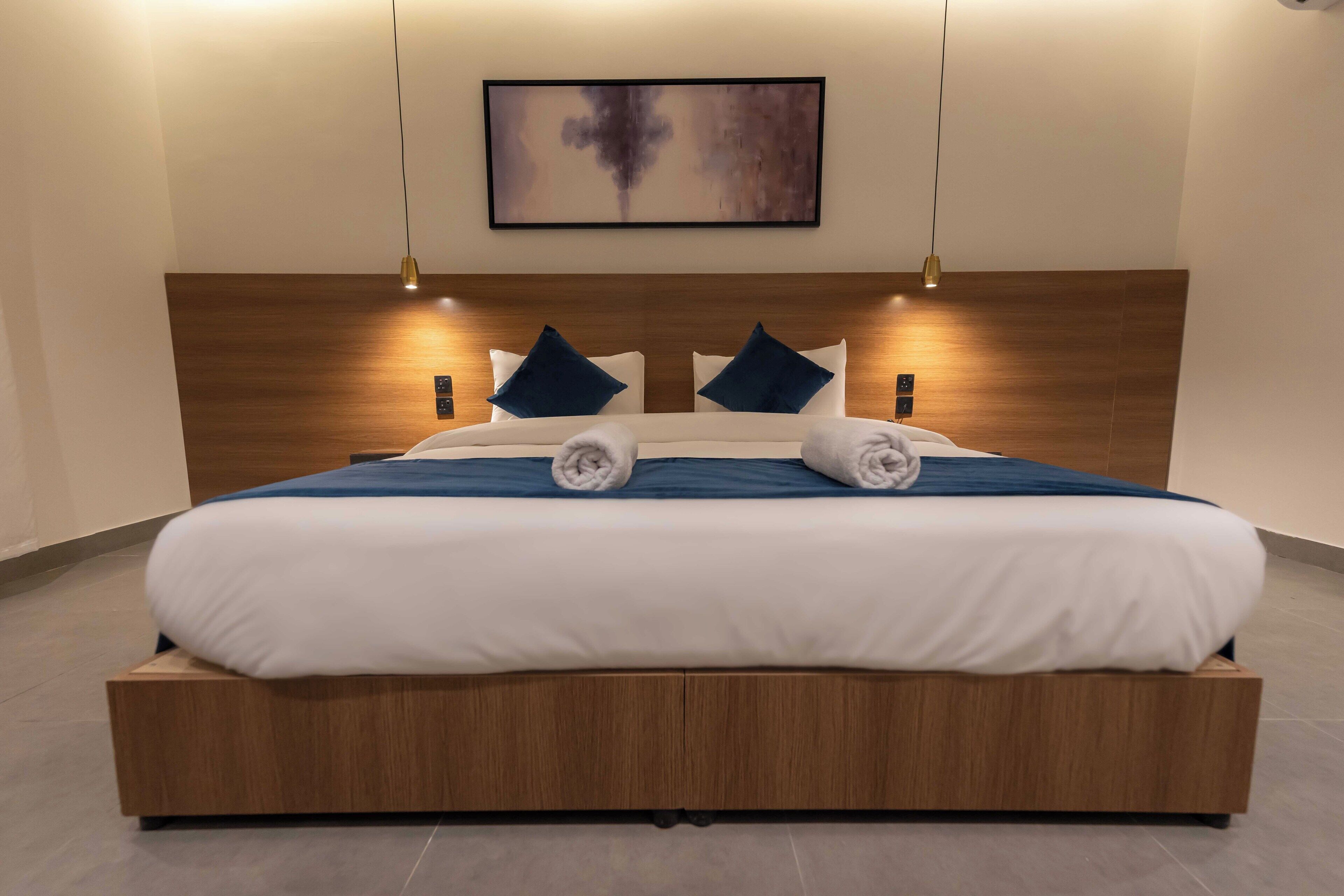 A modern hotel room with a wooden headboard, a large bed with white and blue bedding, two blue pillows, and a wall-mounted abstract painting.