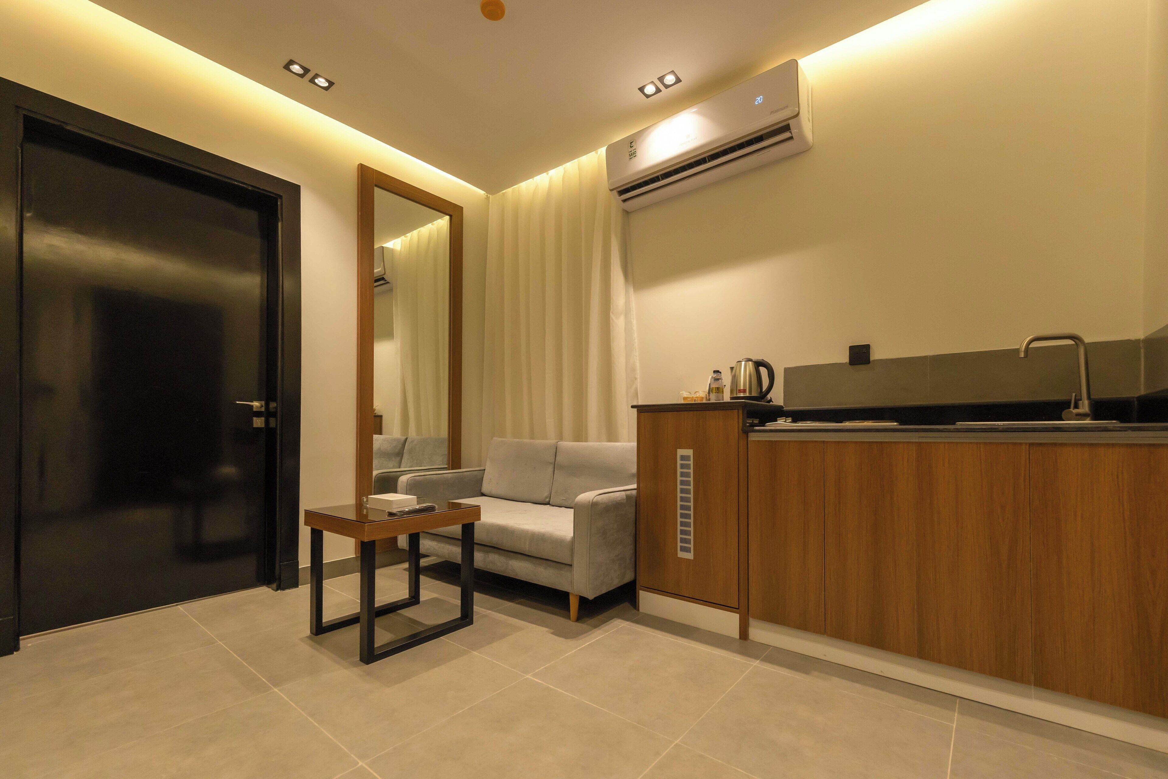 A modern hotel room with a sofa, a small table, a kitchenette with a coffee maker, and an air conditioning unit.