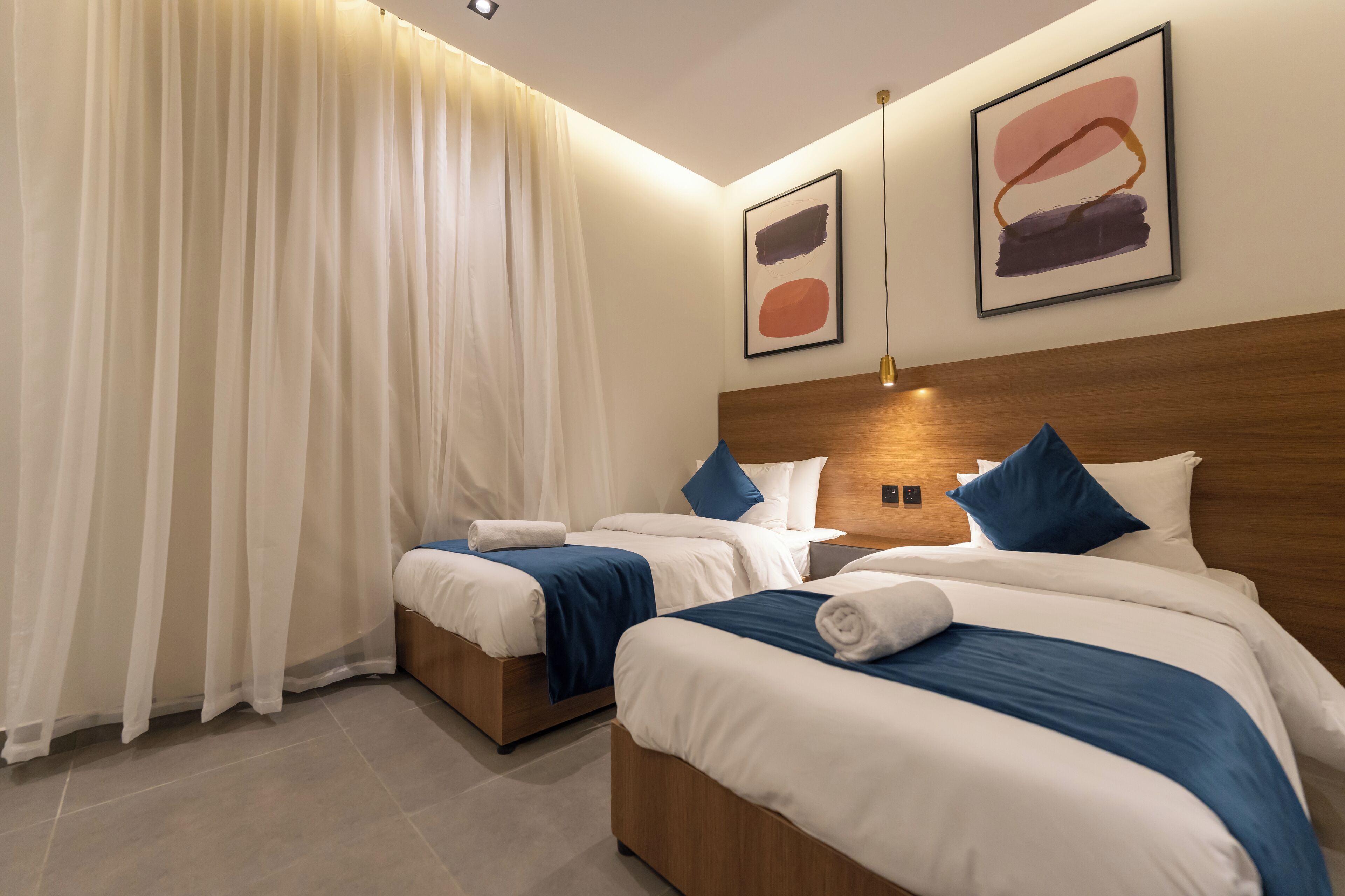 A hotel room with two beds, a wooden headboard, a large window with sheer curtains, and abstract art on the wall.