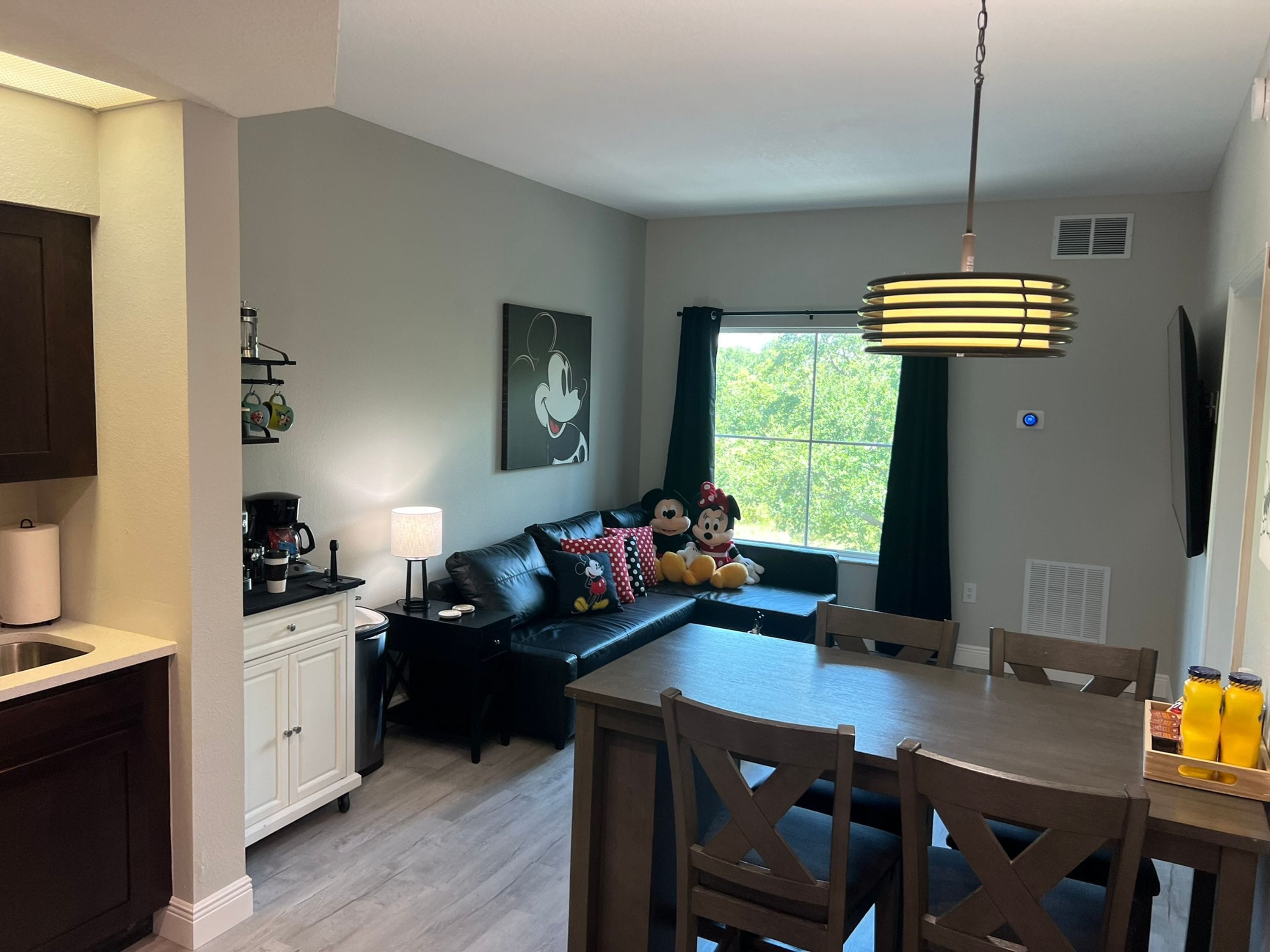 A modern kitchen with dark cabinetry, a dining area with a wooden table and chairs, a black leather sofa with plush toys, and a large window with curtains.